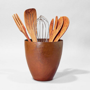 Hand-hammered utensil jar with a wisk and wooden cooking utensils inside centered on a grey background