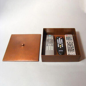 This image shows a hand-hammered remote box. The box is a rectangular shape and the lid comes off using a circular pull. There are three remotes sitting inside this remote box.