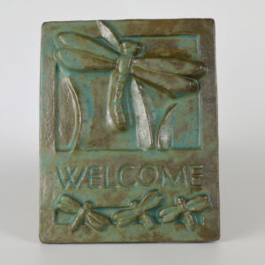 This image shows our dragonfly welcome tile produced by Janet Ontko. The ceramic plaque is glazed in a green glaze that is more green where it is thickest and more brown where it is thin.