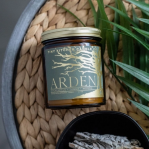 This image shows the candle Arden. It is staged on a braided circular background with a palm leaf to the right of the candle and burning eucalyptus underneath.