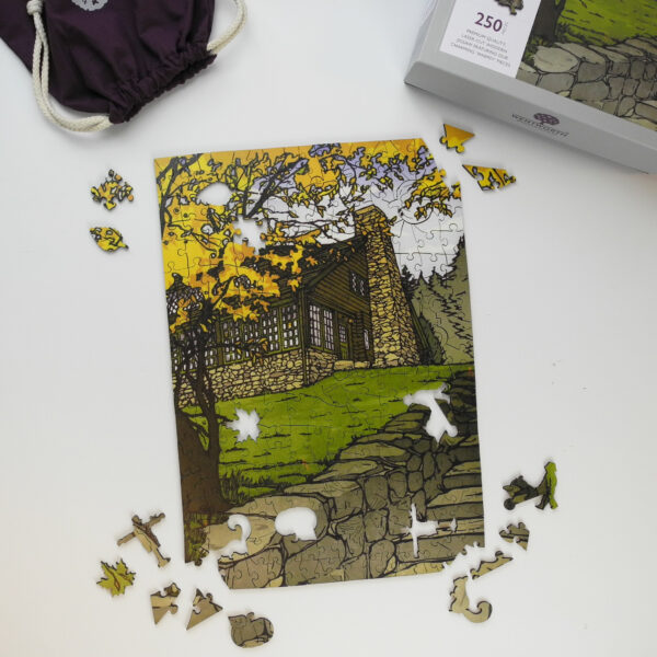 This image shows all the contents you would receive if you purchased a puzzle featuring Yoshiko Yamamoto's print the Log House in Autumn. This image highlights the puzzle, box and purple bag for the puzzle pieces.